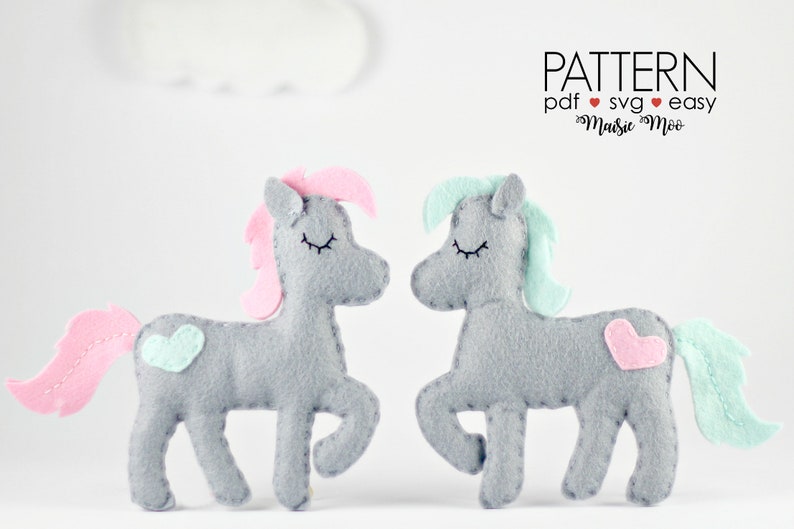 May include: Two gray felt unicorn toys with pink and blue felt manes and tails. The unicorns have closed eyes and pink and blue felt hearts on their sides. The text "PATTERN pdf &hearts; svg &hearts; easy  &hearts;  Maisie Moo" is in the upper right corner of the image.