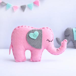 May include: A pink felt elephant plush toy featuring a gray ear patch with a light blue heart accent stitched in the center. The elephant has embroidered closed eyes and visible blanket stitching along the edges of its body and ear.