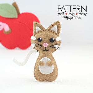 May include: A brown felt cat with white accents, including a white tail, white paws, and white markings on its face. The cat is standing on a white surface. The text "PATTERN pdf ♥ svg ♥ easy Maisie Moo" is visible in the upper right corner of the image.