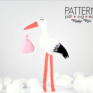 Puede incluir: Una cigüeña de fieltro blanco con una bolsa de fieltro rosa en su pico. La cigüeña tiene patas rojas y alas negras con detalles de purpurina. El fondo es blanco con bolas de algodón.