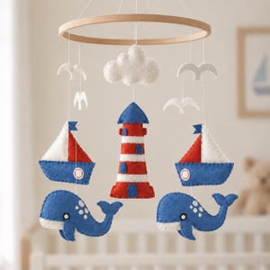 May include: A nautical-themed mobile featuring a circular wooden frame with hanging felt ornaments. The set includes a red and white striped lighthouse, two blue and white sailboats, two blue whales, a white cloud, and four white bird silhouettes.