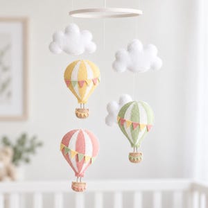 May include: A nursery mobile featuring three felt hot air balloons in yellow, green, and pink, each decorated with small triangular bunting. Two white felt clouds hang alongside the balloons, all suspended from a circular white frame.