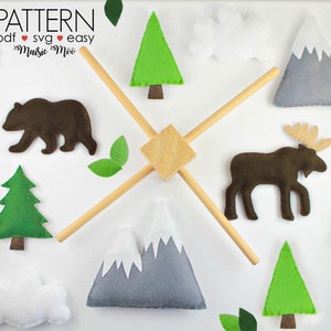 May include: A felt woodland mobile with a bear, moose, trees, mountains, clouds, and leaves. The mobile has a wooden cross-shaped frame and a central wooden cube. The text "PATTERN PDF SVG EASY Maisie Moo" is visible.