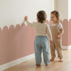 Può includere: Due bambini piccoli sono in piedi vicino a un muro decorato con un bordo a conchiglia rosa polveroso. Un bambino indossa una camicia a maniche lunghe color crema e pantaloni azzurri. L'altro indossa una tuta color cachi. La stanza ha un pavimento in legno chiaro.