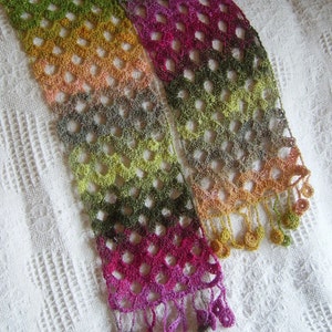 Puede incluir: Una bufanda de ganchillo con un patrón multicolor de verde, amarillo, naranja, rosa y morado. La bufanda tiene flecos en la parte inferior.