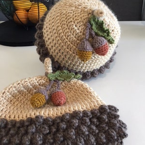 May include: Two handmade crochet hats. The hats are beige with a dark brown textured brim. Each hat is decorated with a cluster of colourful acorns and green leaves. The hats are on a white surface.