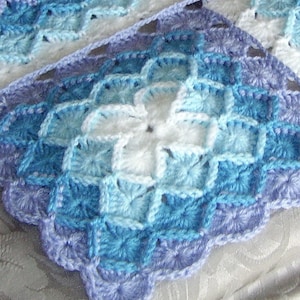 May include: A crocheted blanket in various shades of blue and white. The blanket has a repeating square pattern with a scalloped border. The colour scheme transitions from light blue to a darker blue and white in the centre.