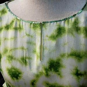 May include: A light-coloured dress with a tie-dye pattern in shades of green. The neckline is trimmed with green sequins. The dress is displayed on a mannequin.