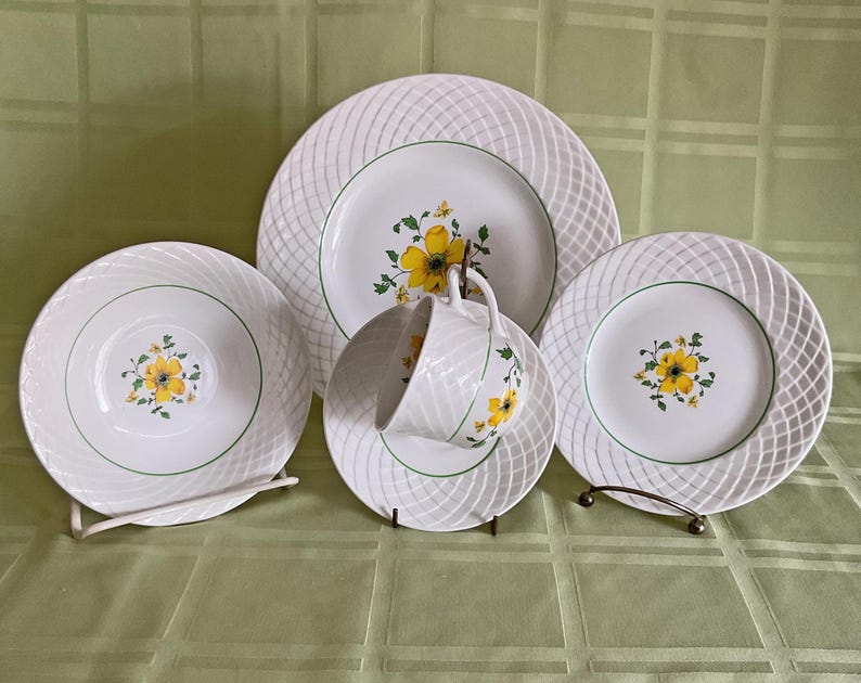 May include: A set of white ceramic dinnerware with a woven pattern and yellow floral design. The set includes a dinner plate, a salad plate, and a saucer.
