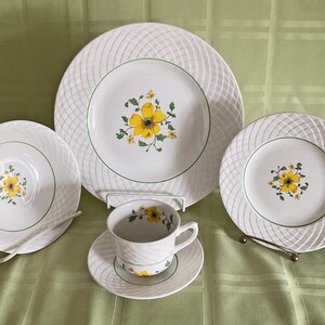 May include: A set of white ceramic dinnerware with a woven pattern and yellow floral accents. The set includes a dinner plate, salad plate, and a teacup and saucer.