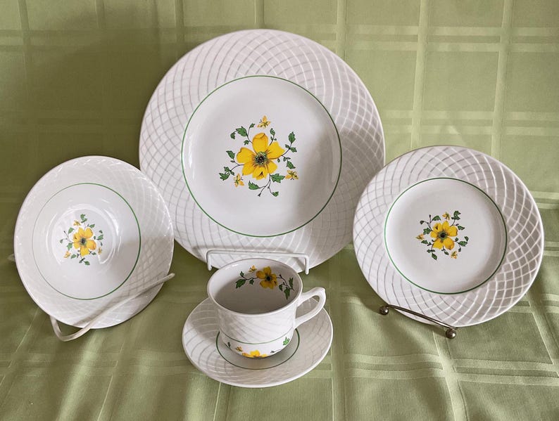 May include: A set of white ceramic dinnerware with a woven pattern and yellow floral accents. The set includes a dinner plate, salad plate, and a teacup and saucer.