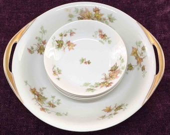 May include: A set of three white porcelain bowls with a gold rim and a floral pattern of green and brown leaves. The bowls are nested inside each other, with the largest bowl having two handles.