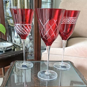 May include: Three ruby red crystal wine glasses with clear stems and bases. Each glass features a distinct etched pattern: one with diagonal swirls, one with a diamond lattice design, and one with a cross-hatch band near the rim. All have clear glass.