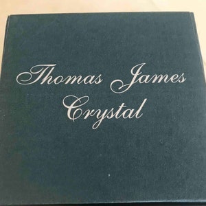 Puede incluir: Una caja azul oscuro con las palabras "Thomas James Crystal" en elegante escritura blanca. La caja tiene una superficie ligeramente texturizada y un dise&ntilde;o simple y cl&aacute;sico, lo que sugiere un producto de alta gama.