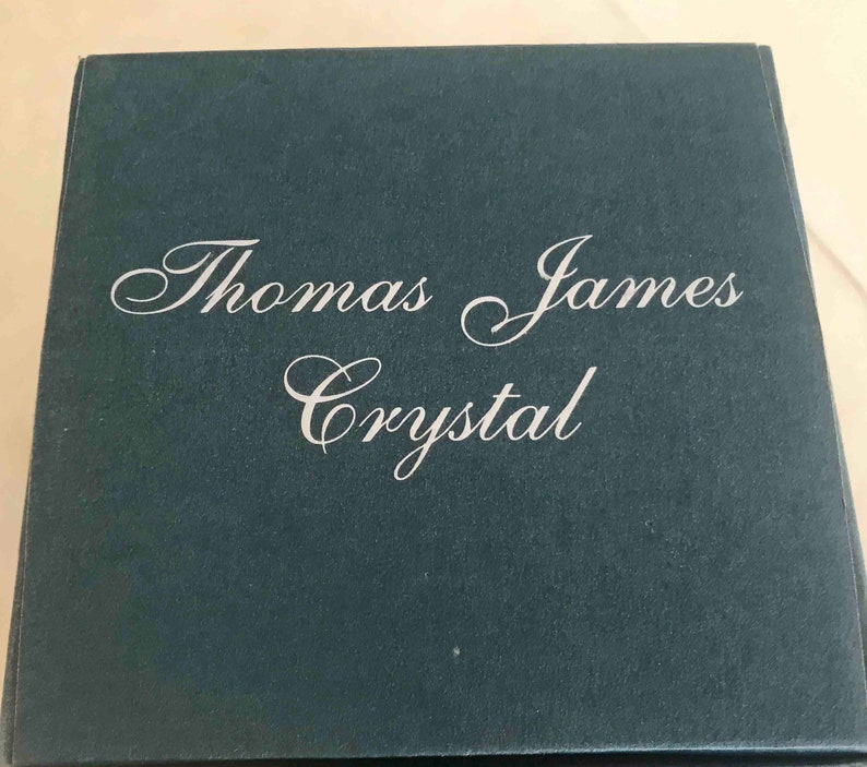 Puede incluir: Una caja azul oscuro con las palabras "Thomas James Crystal" en elegante escritura blanca. La caja tiene una superficie ligeramente texturizada y un dise&ntilde;o simple y cl&aacute;sico, lo que sugiere un producto de alta gama.