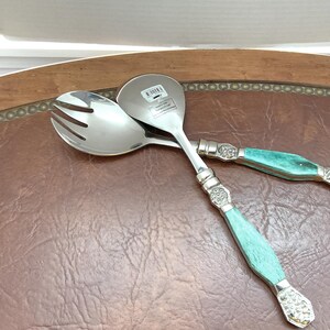 May include: A pair of stainless steel salad servers with turquoise handles. The serving spoon and fork are crossed, showing the silver metal and decorative handles. The handles have a silver band at the top and bottom.