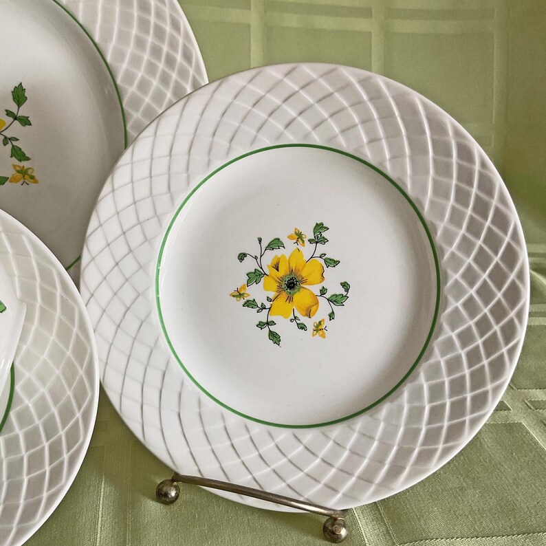 May include: A set of three white ceramic dinner plates with a woven pattern and a green rim. The center of the largest plate features a yellow flower with green leaves.