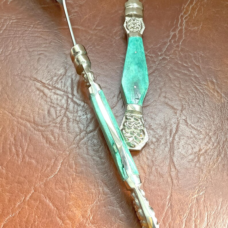 May include: Close-up of two silver serving utensils with turquoise handles. The handles have a decorative design and are accented with silver. The utensils are on a brown leather surface.