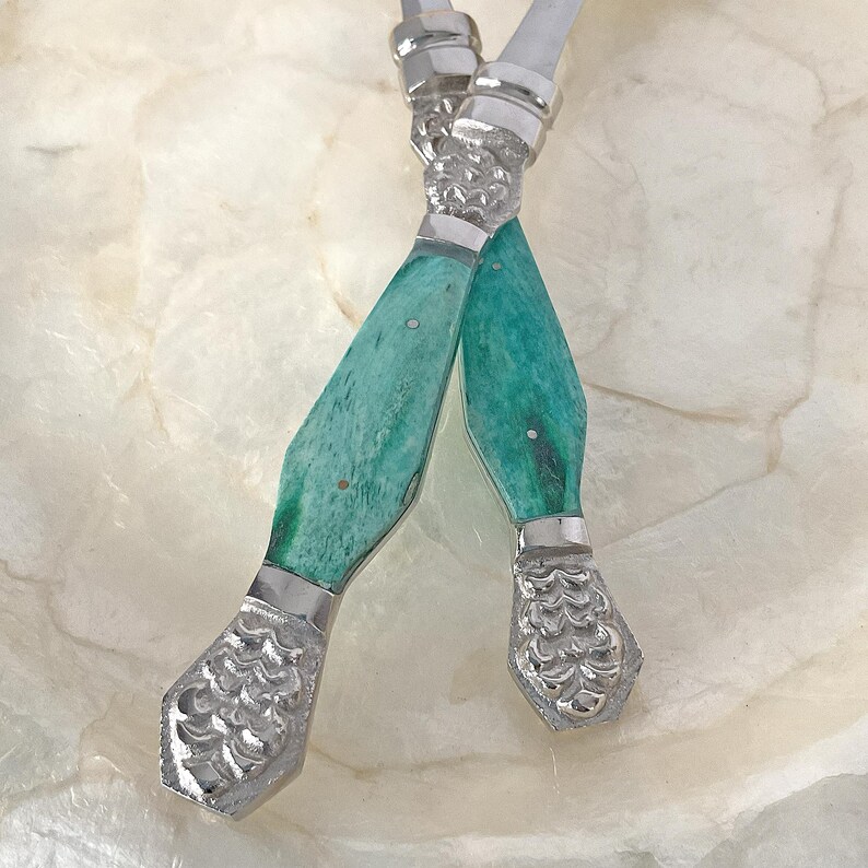 May include: Two crossed silver butter knives with turquoise handles. The handles have a marbled effect and are accented with silver bands. The ends of the knives have a decorative, textured design.