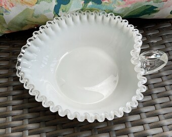 May include: A white, ruffled-edge glass bowl with a clear glass handle. The bowl has a smooth, glossy interior and a delicate, wavy rim. The bowl is likely vintage and suitable for serving.