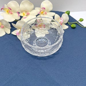 May include: A clear, cut-glass bowl with a diamond pattern and a decorative base. The bowl is set on a blue surface, with white and pink orchids in the background. The bowl is likely used for serving or display.