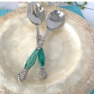 May include: Salad servers with silver heads and teal handles. The handles have a textured silver band at the top and bottom. The servers are crossed over a decorative, off-white plate with a mosaic-like border. The plate is approximately 25 cm in diameter.