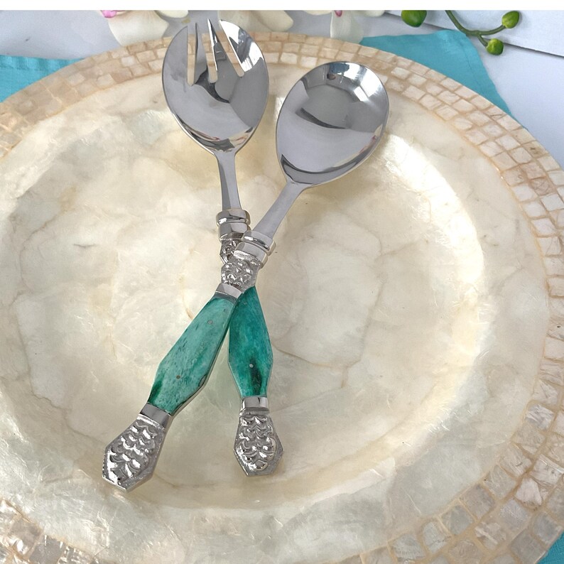 May include: Salad servers with silver heads and teal handles. The handles have a textured silver band at the top and bottom. The servers are crossed over a decorative, off-white plate with a mosaic-like border. The plate is approximately 25 cm in diameter.