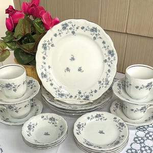 May include: A white porcelain dinnerware set featuring a delicate blue floral pattern. The collection includes dinner plates, saucers, cups, and small bowls, all decorated with small blue rose clusters and scalloped edges on the plates.