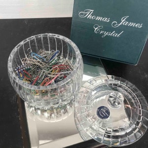 Puede incluir: Un cuenco de cristal transparente y redondo lleno de clips de papel de colores, con una tapa a juego. El cuenco y la tapa tienen un patr&oacute;n de rejilla. Una caja verde con las palabras "Thomas James Crystal" est&aacute; en el fondo.