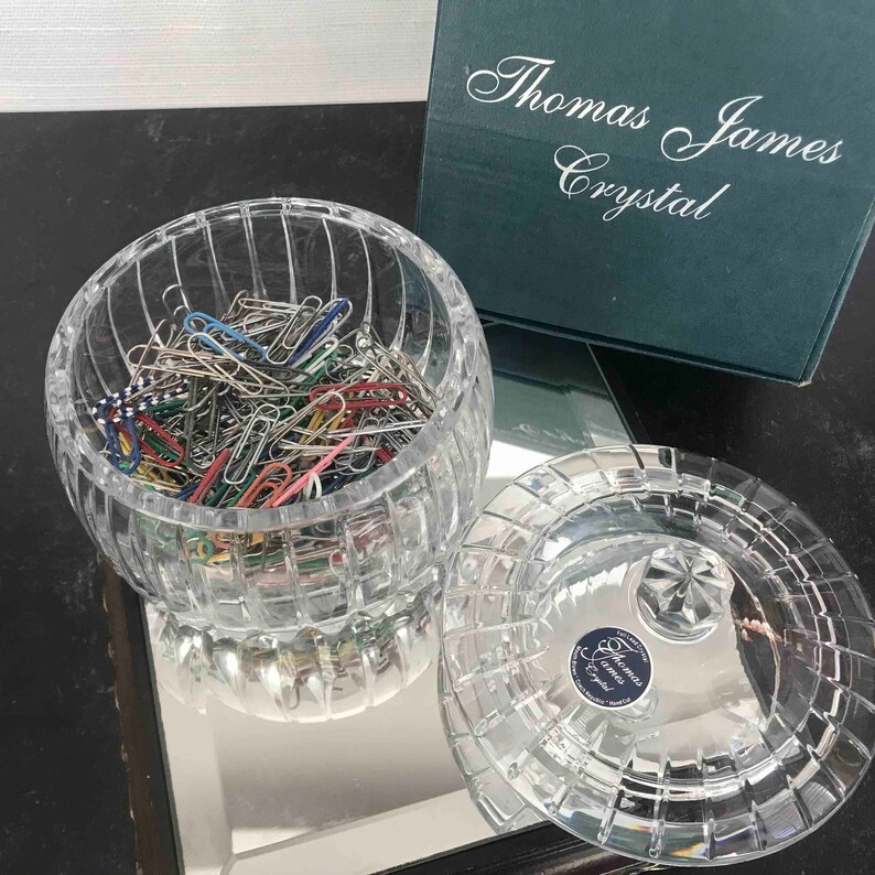 Puede incluir: Un cuenco de cristal transparente y redondo lleno de clips de papel de colores, con una tapa a juego. El cuenco y la tapa tienen un patr&oacute;n de rejilla. Una caja verde con las palabras "Thomas James Crystal" est&aacute; en el fondo.