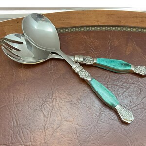 May include: Two salad servers with stainless steel heads and turquoise handles. The handles feature decorative silver-coloured metal detailing. One server is a spoon, the other a fork.