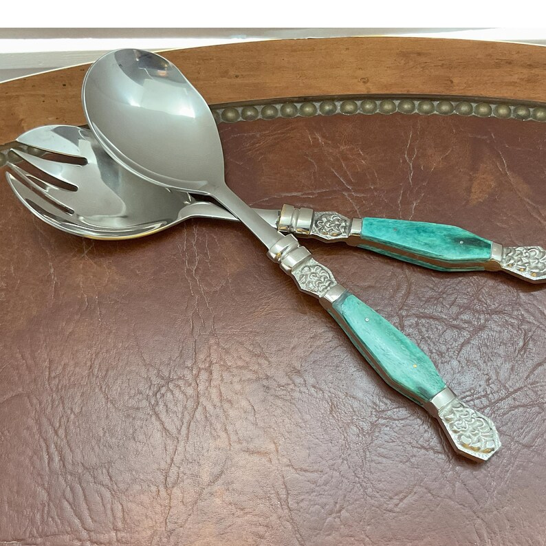 May include: Two salad servers with stainless steel heads and turquoise handles. The handles feature decorative silver-coloured metal detailing. One server is a spoon, the other a fork.