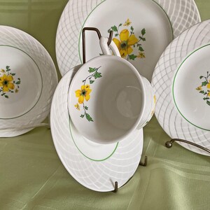 May include: A white teacup and saucer with a green and yellow floral design. The saucer has a woven pattern and a green trim. The teacup has a yellow flower with a green stem and leaves.