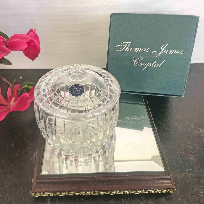 Puede incluir: Un tarro de cristal transparente con tapa, colocado sobre una superficie espejada. El tarro tiene un patr&oacute;n tallado en forma de diamante y una peque&ntilde;a etiqueta azul y blanca. Una caja verde con "Thomas James Crystal" est&aacute; en el fondo.
