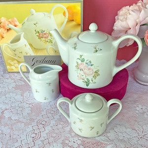 May include: A white porcelain tea set by Gorham featuring a teapot, a creamer, and a lidded sugar bowl. Each piece is decorated with a delicate floral pattern of pink roses and green leaves. The set is displayed with its original product box.