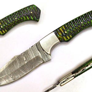 Può includere: Un coltello fatto a mano con una lama a fantasia e un manico verde e giallo. La lama ha un design unico e strutturato. Il manico è un mix di verde e giallo con piccoli accenti dorati. Il coltello è mostrato da più angolazioni.