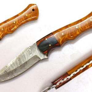 Può includere: Coltello da caccia fatto a mano con lama in acciaio Damasco e manico in legno. Il manico è di colore marrone caldo con un motivo marmorizzato e rivetti in ottone. Il coltello è mostrato con due pezzi di manico aggiuntivi.