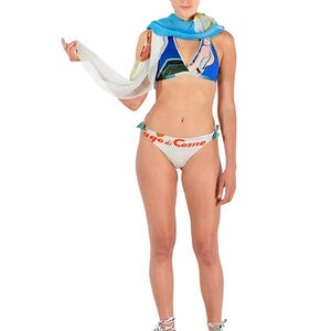 Puede incluir: Una mujer lleva puesto un bikini blanco con el texto "Lago di Como" impreso. También lleva un bikini azul de halter y una bufanda blanca.