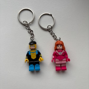 Peut inclure: Deux porte-clés avec des figurines en plastique. L'une porte une combinaison bleue et jaune avec des détails noirs et des lunettes. L'autre a des cheveux orange et une combinaison rose avec un emblème circulaire sur la poitrine. Anneaux en métal argenté.