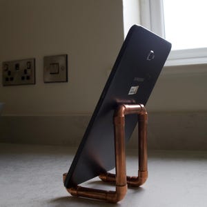 May include: A dark grey Samsung tablet resting on a copper pipe stand. The stand is constructed from interconnected copper pipes and fittings, forming a sturdy base and a vertical support frame that holds the tablet at an angled position.