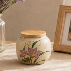 Op de afbeelding: Een ronde keramische pot met een lichtcrème gespikkelde afwerking en een plat houten deksel. De pot is voorzien van een handgeschilderd ontwerp van paarse bloemen met groene bladeren en stengels. De pot heeft een bolvormig lichaam en een brede opening.