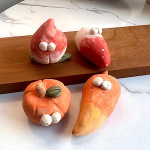 May include: Four whimsical ceramic fruit sculptures: a peach, a strawberry, a pumpkin, and a carrot. Each piece is painted with vibrant colors and features two small, white, googly eyes. The sculptures are arranged on a wooden board.