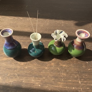 Puede incluir: Cuatro pequeños jarrones de cerámica hechos a mano en tonos verdes, turquesas y morados. Un jarrón contiene varillas de incienso. Los jarrones tienen un acabado brillante y formas únicas, uno de ellos con la parte superior con volantes. Están sobre una superficie de madera.