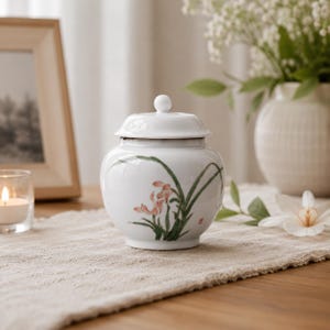 Może przedstawiać: Biały ceramiczny słoik z pokrywką, ozdobiony ręcznie malowanym motywem botanicznym przedstawiającym zielone liście i delikatne różowe kwiaty. Słoik ma zaokrąglony korpus i mały uchwyt na pokrywce, wykończony gładką, błyszczącą powierzchnią.
