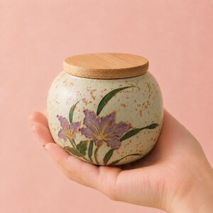 May include: A small, round ceramic jar with a speckled cream-colored finish and a light brown wooden lid. The jar features a hand-painted design of purple flowers with green leaves and stems, showcasing a textured surface and a rounded shape.