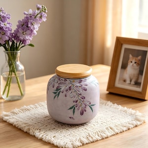 May include: A lavender ceramic jar with a wooden lid, adorned with purple floral designs and green leaves. The jar sits on a cream-colored, fringed placemat. A vase of purple flowers and a framed cat photo are in the background.