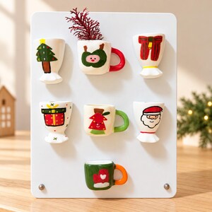以下が含まれることがあります： 白い長方形のマグネットボードに、クリスマスをテーマにした7つの陶器製マグネットが並んでいます。マグネットには、クリスマスツリー、プレゼント、サンタクロースの顔、赤いリボン、そしてお祝いのモチーフが描かれた3つの小さなマグカップが手描きされています。