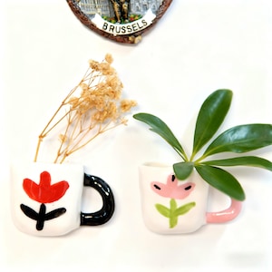 Könnte beinhalten: Zwei kleine, weiße Keramikbecher mit Blumendesigns. Einer hat eine rote Tulpe mit schwarzem Stiel und Griff, der andere eine rosa Tulpe mit grünem Stiel und rosa Griff. Getrocknete Blumen und Grünzeug sind in den Bechern.