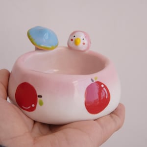 May include: A small, round, pink ceramic bowl with a cartoon bird and mushroom design. The bowl features red apple illustrations and is held in a hand. The bird has a yellow beak and the mushroom is blue with a yellow rim.