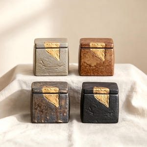 May include: Four square ceramic boxes with lids. Each box has a different colour: grey, brown, dark brown, and black. Each lid features a gold leaf design. The boxes are arranged on a white surface.